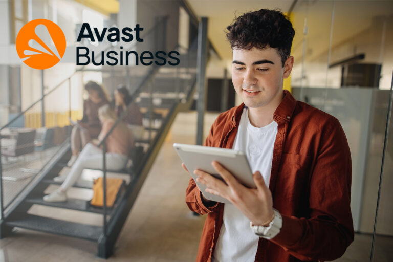 Junger Mann in einer Art Aula oder Schul-Bibliothek stehend den Blick auf sein Tablet gerichtet, im Hintergrund zwei Schülerinnen auf einer Treppe sitzend und ein Avast-Logo