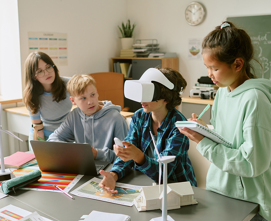 Klassenzimmer der Zukunft - Symbolbild - Gruppe Jugendlicher, die an einem Wissenschaftsprojekt zusammenarbeiten, ein Junge mit einem Virtual-Reality-Headset, während ein Mädchen Notizen macht, Klassenkameraden beobachten und diskutieren über Modelle erneuerbarer Energien