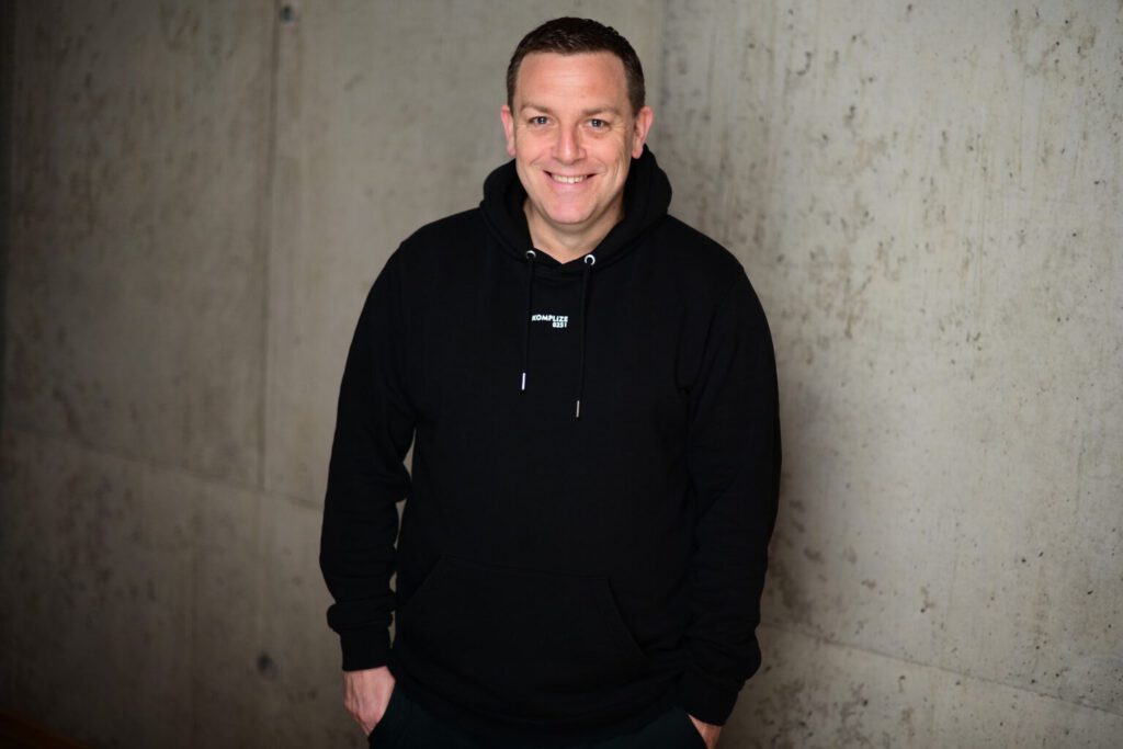 Porträt eines lächelnden Mannes (Andreas Hofmann, Beitragsautor) im schwarzen Hoodie vor einer modernen Betonwand.