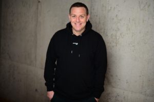 Porträt eines lächelnden Mannes (Andreas Hofmann, Beitragsautor) im schwarzen Hoodie vor einer modernen Betonwand.