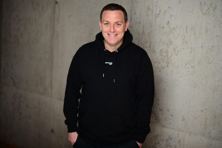 Porträt eines lächelnden Mannes (Andreas Hofmann, Beitragsautor) im schwarzen Hoodie vor einer modernen Betonwand.