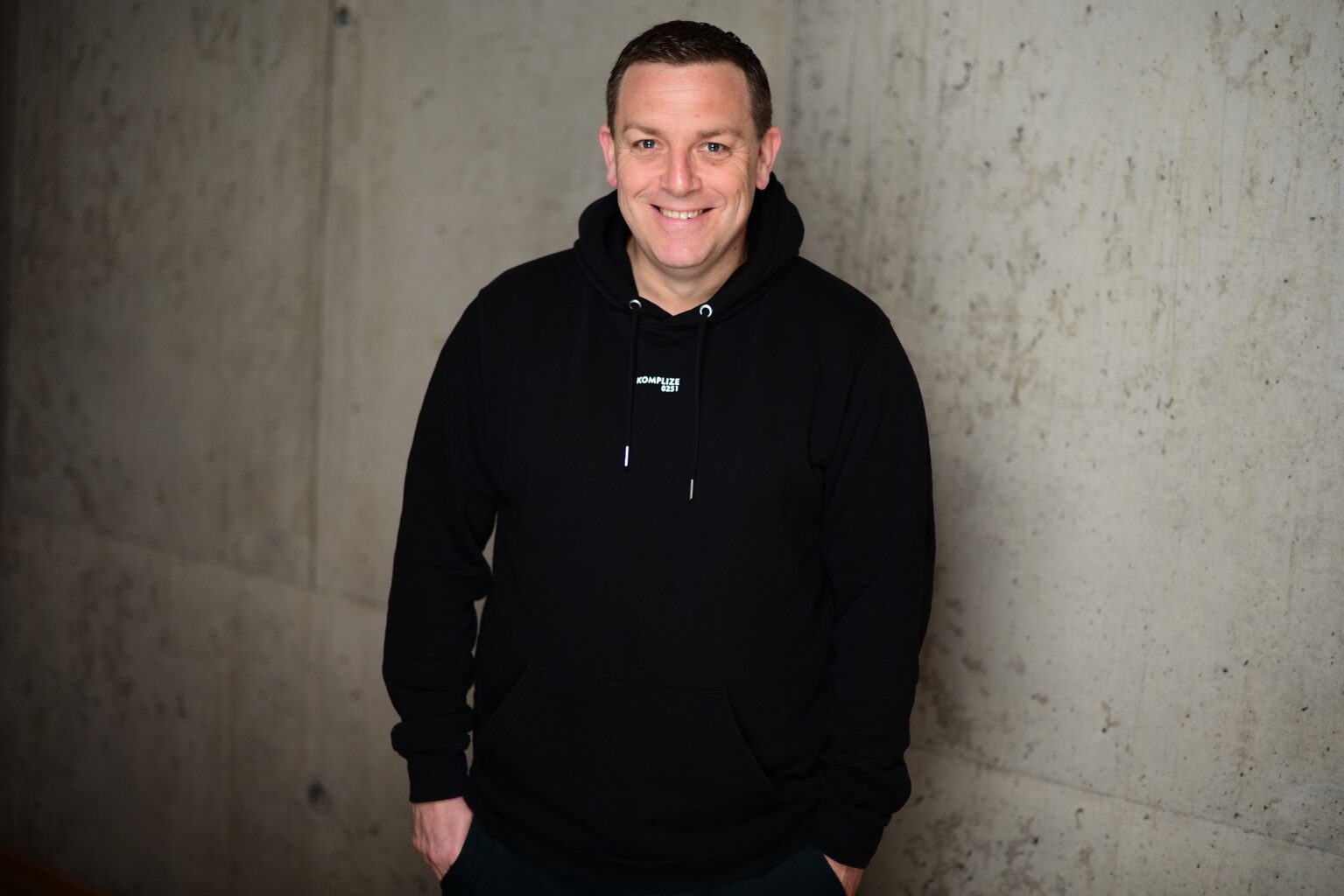 Porträt eines lächelnden Mannes (Andreas Hofmann, Beitragsautor) im schwarzen Hoodie vor einer modernen Betonwand.
